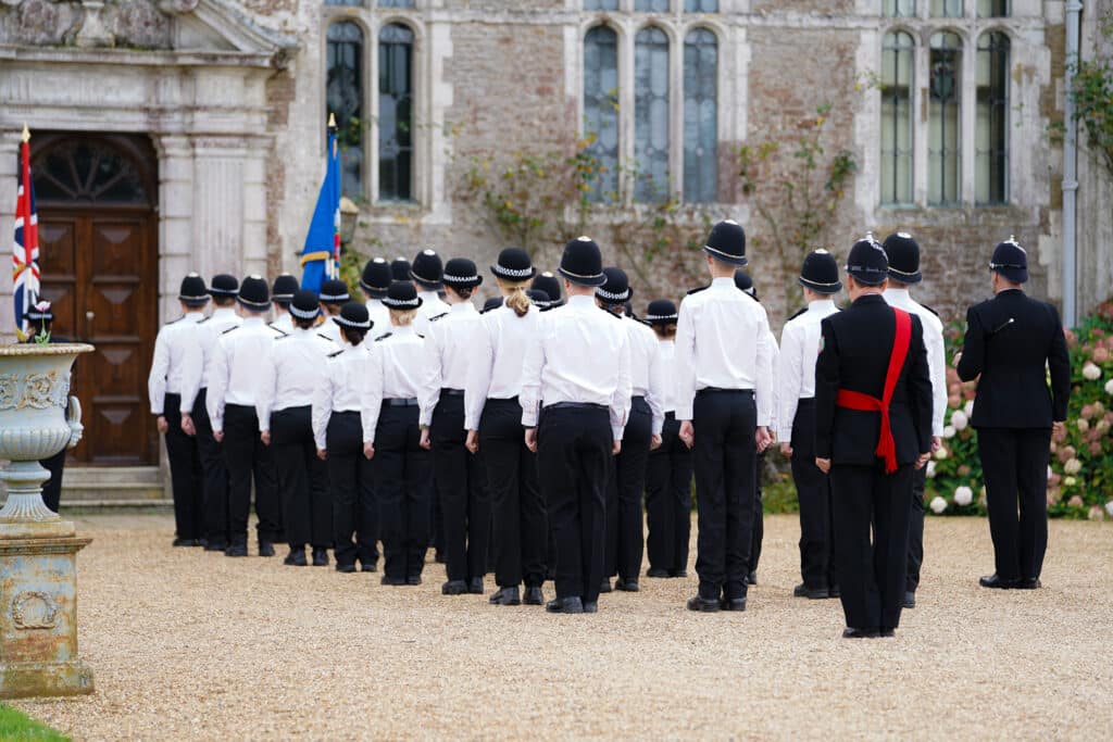 The image shows a large group of officers in white shirts and hats standing with their backs to the camera. In front of the group is a large property, which is Loseley House in Guildford. There are also two flags in front of the group. The image shows a 'passing out parade' for new Surrey Police officers.