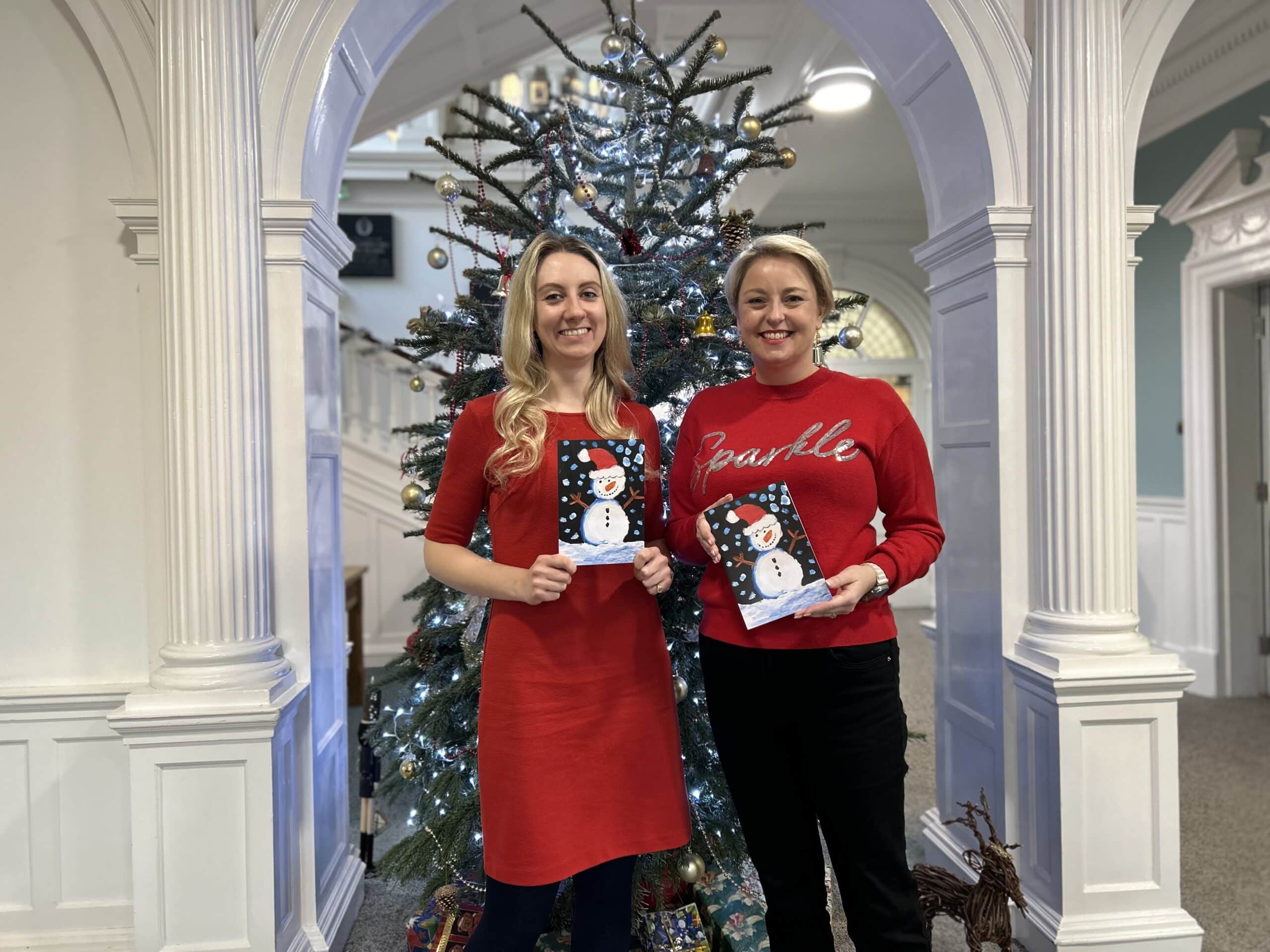 On left, Deputy Police and Crime Commissioner Ellie Vesey-Thompson, and on right, Police and Crime Commissioner Lisa Townsend. Both are standing in front of a Christmas tree and wearing red. They are holding up a Christmas card with a hand-drawn snowman picture on the front.