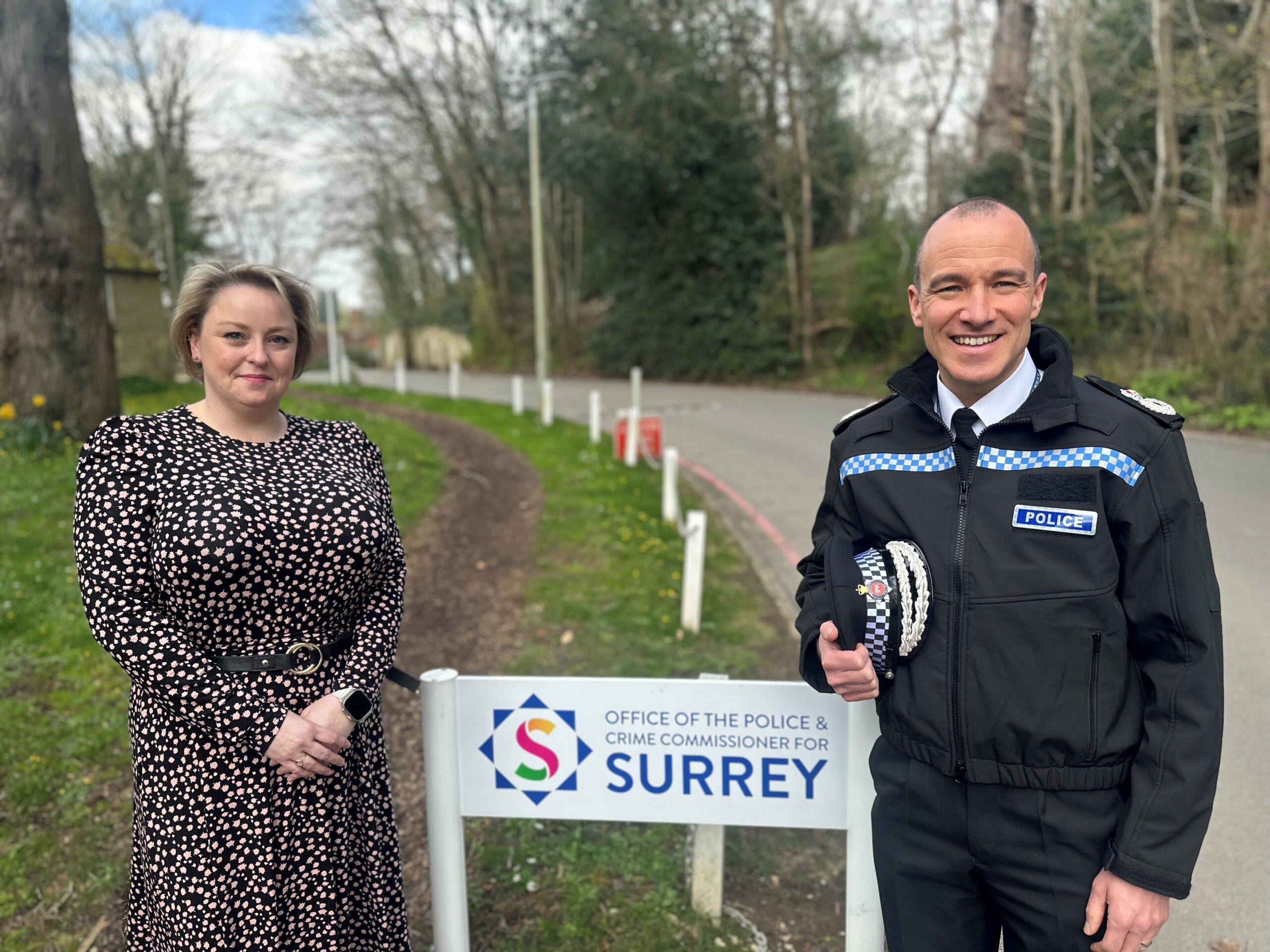 Commissioner Lisa Townsend wears a long-sleeved dress, and has her hands clasped in front of her. Chief Constable Tim De Meyer stands to Lisa's left. He is wearing his uniform and smiling at the camera. He holds his police hat in his right hand. Between Lisa and Tim is a colourful waist-height sign that reads 'Office of the Police and Crime Commissioner for Surrey'