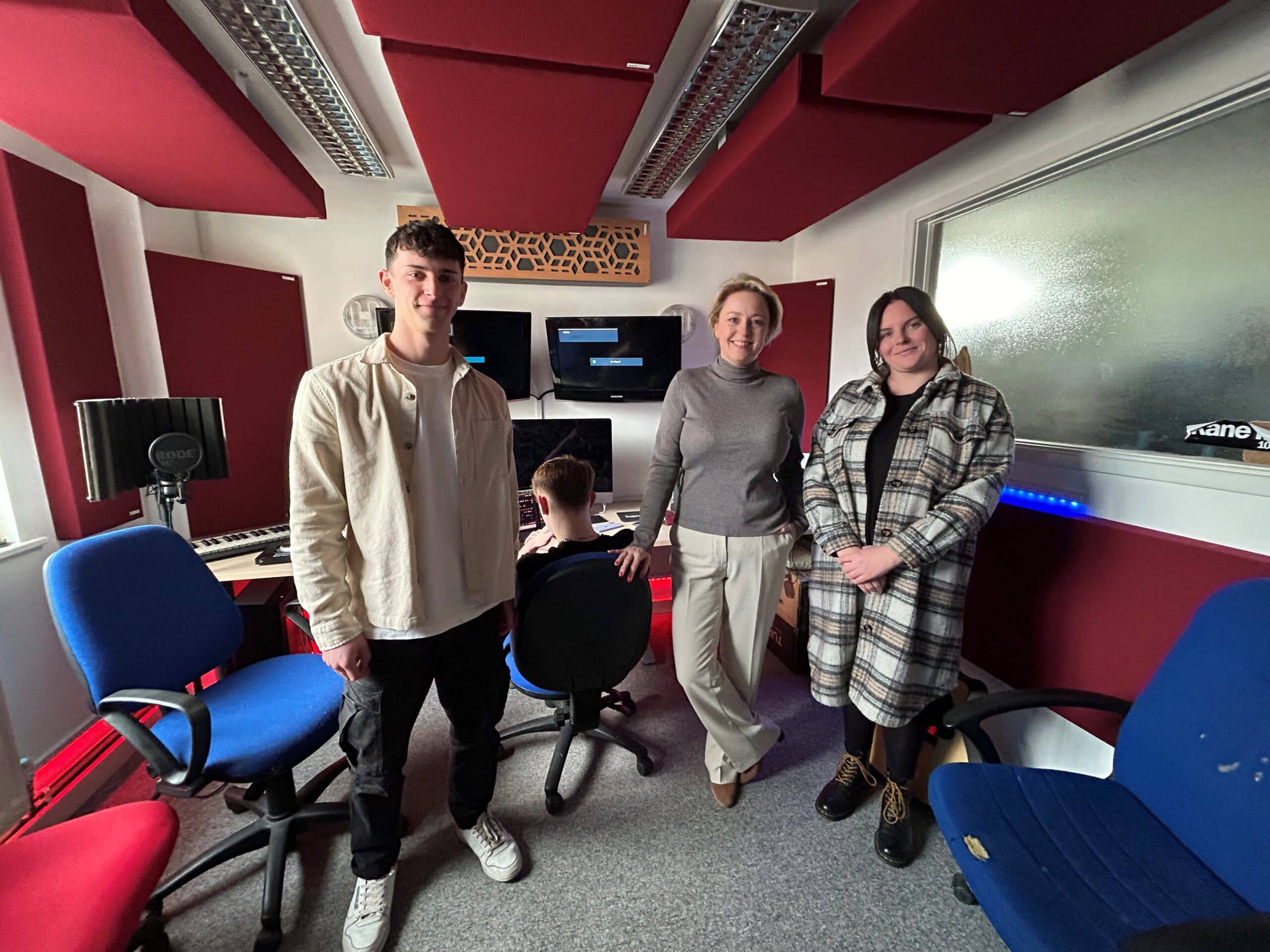 Three people stand in a studio. On the left, a man stands with his hands by his sides. Sitting to his left is a young person with his back to the camera. Next, and in the middle of the standing group, is Police and Crime Commissioner Lisa Townsend, who has her hand on the back of a chair. A woman stands with her hands clasped to Lisa's left hand side.