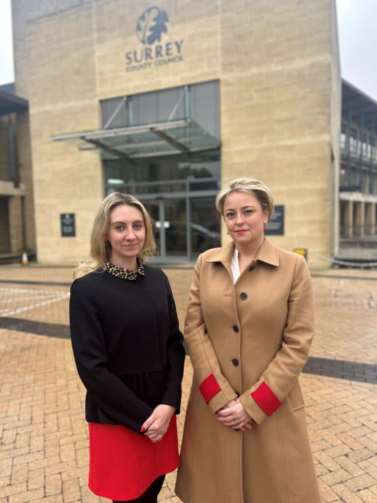 Deputy Police and Crime Commissioner Ellie Vesey-Thompson and Police and Crime Commissioner Lisa Townsend stand next to each other in front of a building with a Surrey County Council sign on it. Both women have their hands clasped in front of them. Ellie, standing on the left, wears a black jumper and red skirt, while Lisa wears a tan coat with red cuffs. Both women are looking straight at the camera.