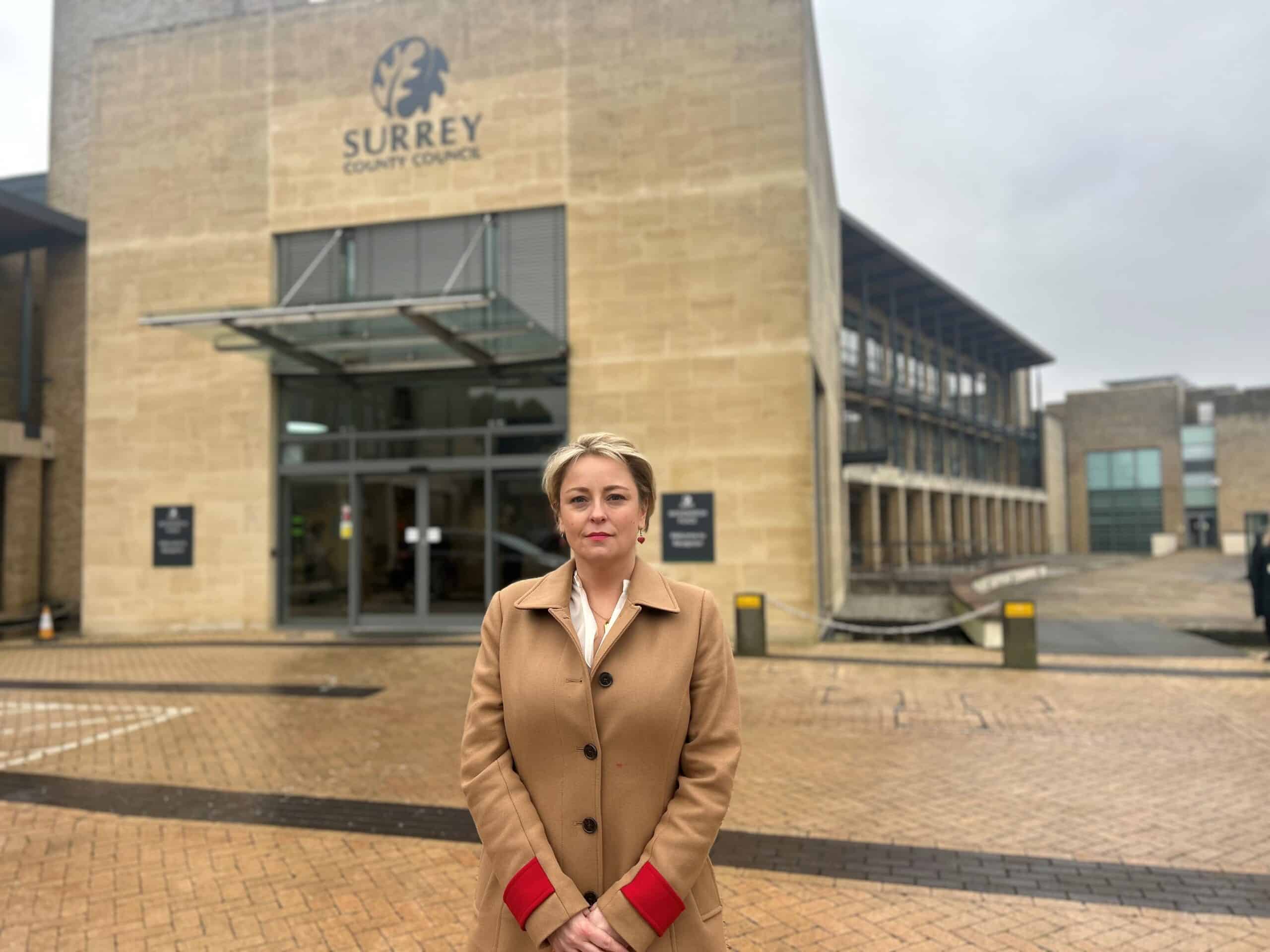 Lisa Townsend, Surrey's Police and Crime Commissioner, stands in front of a large building with a Surrey County Council sign on it. She wears a tan coat with red sleeves. Her hands are clasped in front of her and she is looking straight ahead at the camera