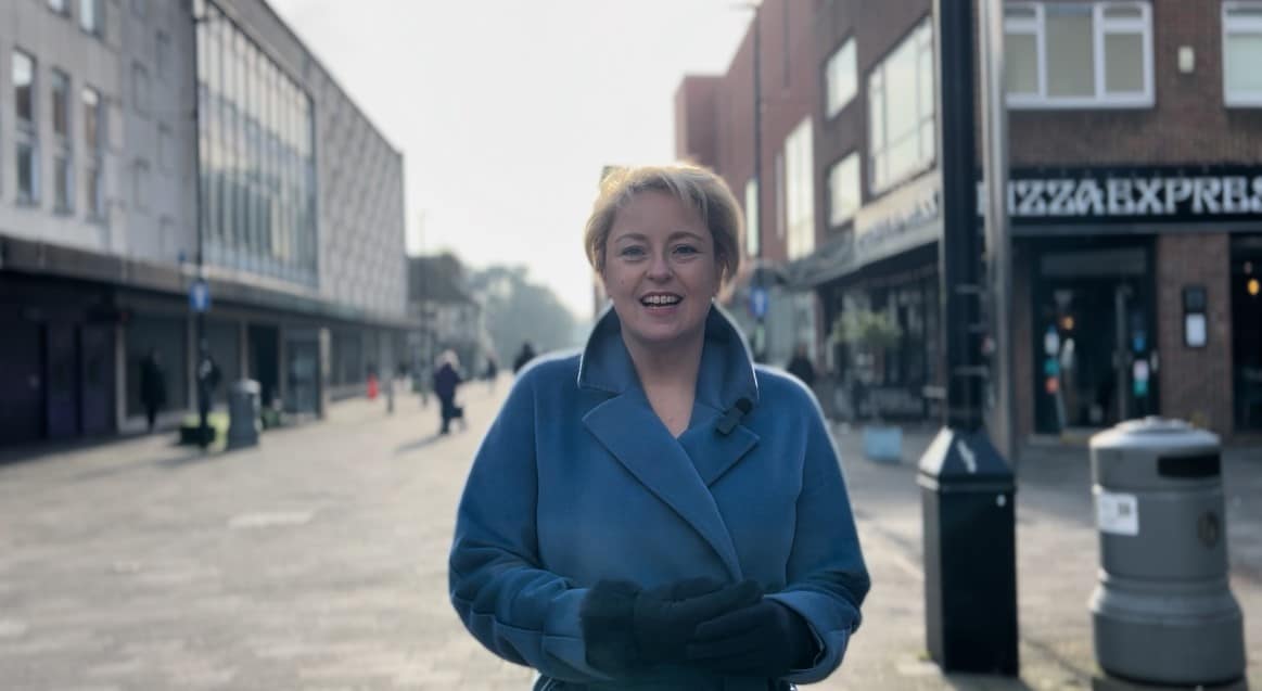 Surrey's Police and Crime Commissioner, Lisa Townsend, stands in a high street. Behind her are shops and people walking. She wears a blue coat with its collar upturned. Her blonde hair is loose and she is smiling.